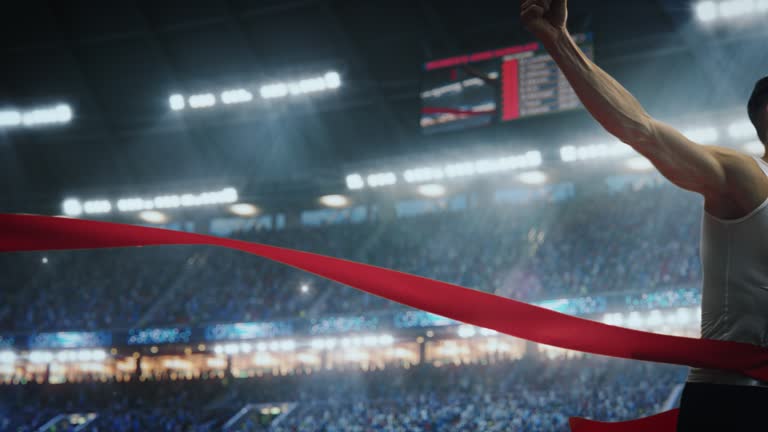 Fit Athlete Finishing a Sprint Run at a Crowded Arena with Cheering Spectators. Young Man Crossing the Finish Line with a Red Ribbon. Cinematic Super Slow Motion Sports Footage