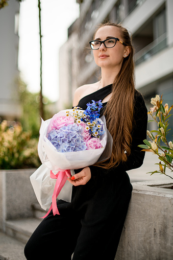 Dreamy brunette girl in black outfit holding huge bouquet of different fresh flowers in wrapping paper outdoors. Variety of delicate flowers in woman's hands. Blurred background