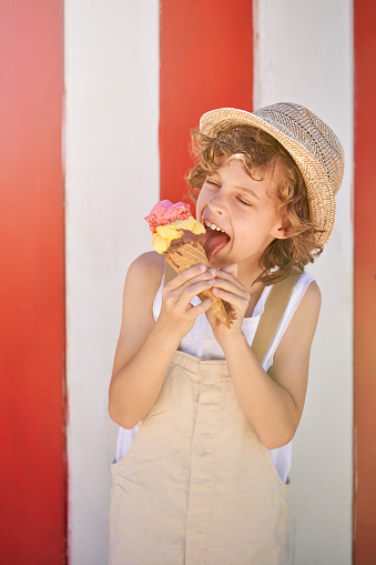 Happy child in straw hat and overall closing eyes in joy while licking sweet ice cream in waffle cone against red and white striped background