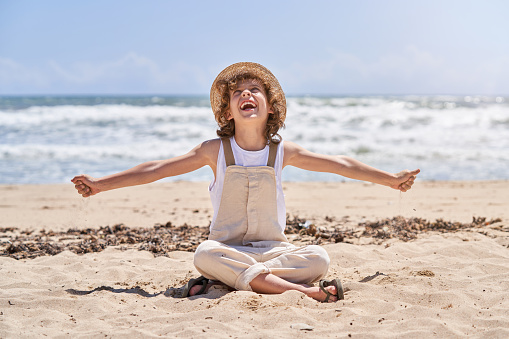 Ground level of full body happy kid in straw hat and jumpsuit pouring sand while playing on seashore and looking up