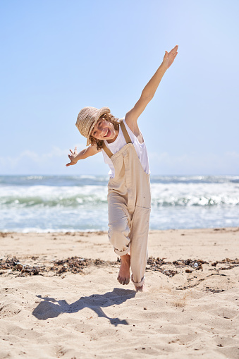 Full body of happy barefoot kid in straw hat running and stretching arms like wings of plane while enjoying summer holidays at seaside