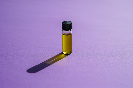 Small bottle of essential oil standing upright on a bright purple background with light shining through it