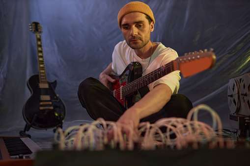 Medium full shot of male musician in yellow beanie sitting on studio floor adjusting musical equipment for electric guitar recording