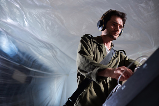 Medium shot with low angle view of adult male composer with stubble wearing khaki shirt and headphones looking down at studio equipment