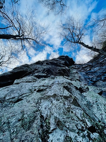 View of cold rock cliff face from below. Bare winter trees and cloudy winter sky loom above.