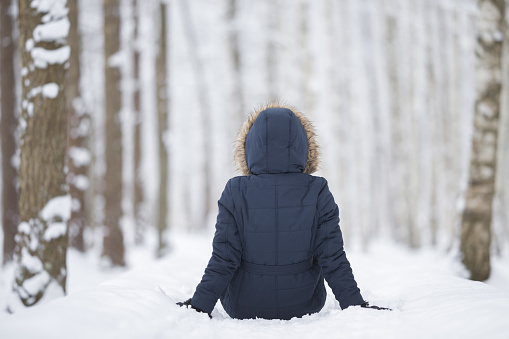 Young adult woman sitting on fresh white snow at birch tree forest. Looking far away. Spending time alone in beautiful cold winter day. Back view. Thinking about life. Peaceful atmosphere in nature.