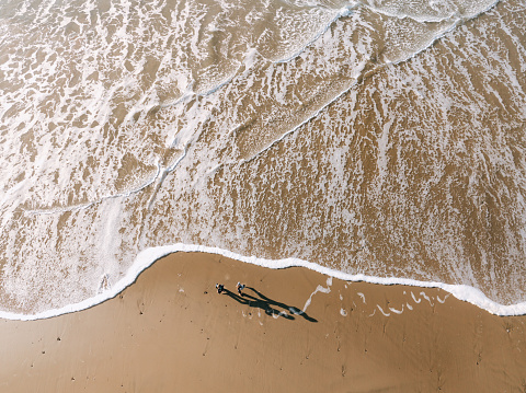 Aerial drone footage from the coast of the Pacific Northwest, featuring Oregons beautiful rugged terrain and blue ocean waves. Children runs from the waves as they foam onto the beach of the Pacific Ocean in Oregon state.  Shot directly above, looking down.