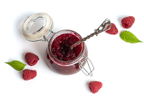Homemade raspberry jam in a small glass jar and fresh fruit on a white background