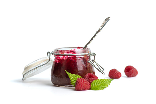 Homemade raspberry jam in a small glass jar and fresh fruit on a white background