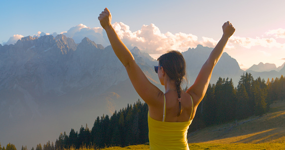CLOSE UP: Sporty lady with raised arms celebrates reaching scenic mountain peak. Young woman feels victorious when she reaches the top, where a picturesque view of high alpine mountains awaits her.