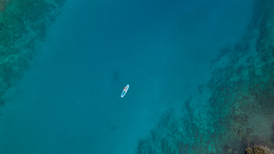 AERIAL TOP DOWN: Young lady stand up paddling in stunning clear blue sea water. A meditative and peaceful water sports activity to explore and admire the surrounding seashore and pristine nature.