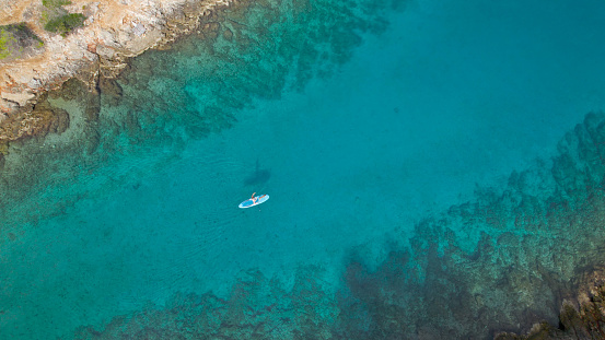 AERIAL TOP DOWN: Female stand up paddler paddling in amazing clear blue sea. She is on active holidays with her doggy at a stunning tourist destination of the island of Hvar with many hidden bays