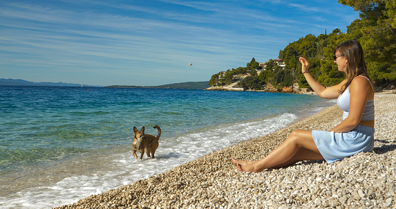 Pretty holiday maker on a pebble seashore throwing stones to her playful dog. She is on summer holidays at the seaside with her dog. They enjoy playing on a beautiful beach by the blue Adriatic Sea.