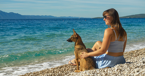 Cute little dog and its pretty owner sit at pebble shore and watch wavy blue sea. Young tourist and her dog travel across Dalmatia and explore stunning and pet friendly beaches by crystal Adriatic Sea