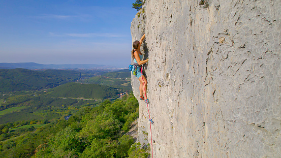 AERIAL: Sporty woman climbing a limestone wall above amazing Karst landscape. Female lead climber is making good progress on a sharp end of rope. Stunning views during challenging climb at Karst Edge.