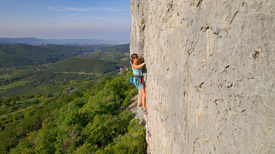 AERIAL: Incredible view from a rocky climbing wall at picturesque Karst Edge. Fearless female climber is successfully overcoming difficulties while climbing challenging route in a limestone wall.