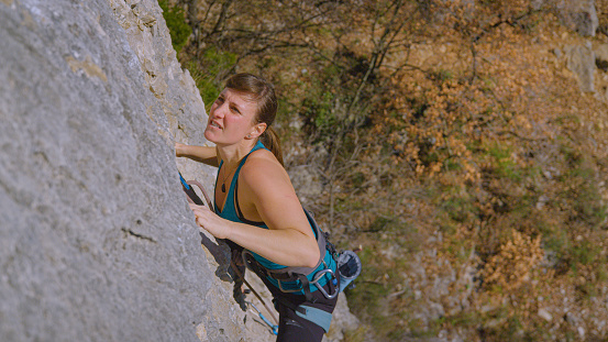 CLOSE UP, DOF: Focused rock climber during ascent of a difficult climbing route. Young female climber advancing in the middle of steep limestone wall. Challenging outdoor sport activity in rocky Karst
