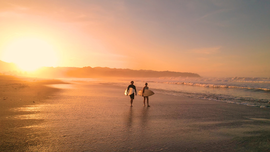 AERIAL: Young couple walking on beautiful sandy beach after sunset surf session. Stunning view of orange glowing sandy beach and rolling ocean waves. Surf couple on a surf trip at exotic Playa Venao.