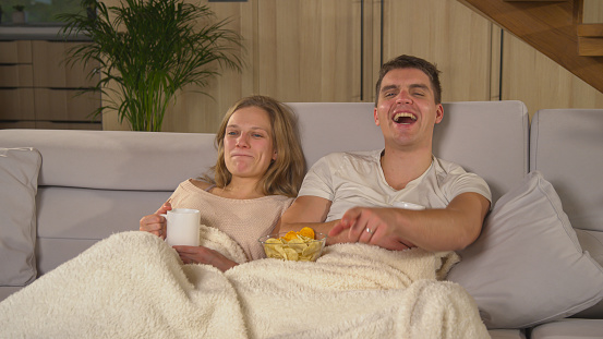 CLOSE UP: Young couple enjoying on comfy sofa and watching entertaining TV show. Married couple under blanket and equipped with tea and snacks for watching movie in living room on a winter evening.