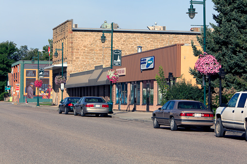 Main street downtown of Pincher Creek in August. Hanging baskets of flowers on light posts. Cars parked. Old historic Sandstone buildings preserved and still used as small local business.