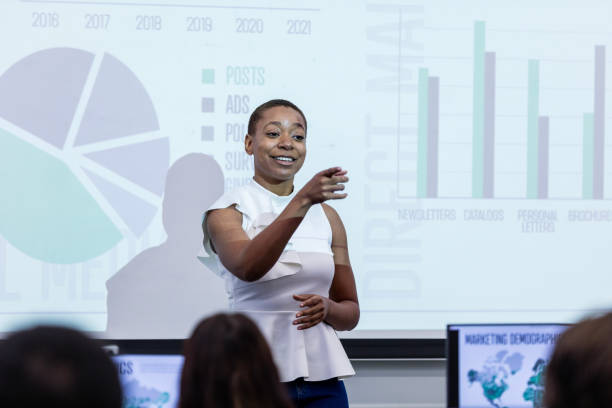 Businesswoman gestures to unseen seminar attendee during lecture The young adult businesswoman gestures to an unseen seminar attendee during her lecture. projector screen classroom stock pictures, royalty-free photos & images