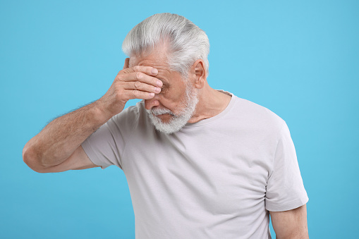Embarrassed senior man covering eyes on light blue background
