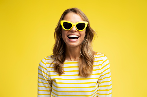 Smiling young woman wearing stylish sunglasses looking at camera isolated on yellow background. Fashion model posing for pictures in studio. Summer, travel, vacation concept