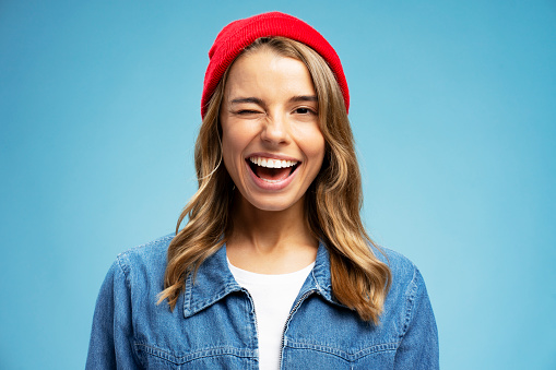 Closeup portrait smiling American woman winks wearing stylish red hat isolated on blue background. Happy modern hipster with stylish hair looking at camera. Positive lifestyle concept