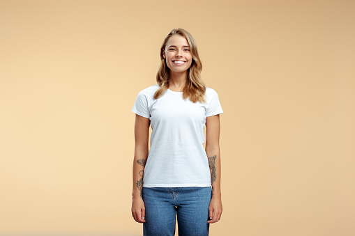 Portrait of smiling beautiful woman with stylish tattoo on arms wearing white t shirt pointing looking at camera isolated on beige background, copy space. Mockup