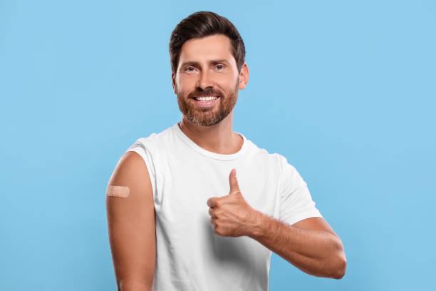 istock Man with sticking plaster on arm after vaccination showing thumbs up against light blue background 1893573311