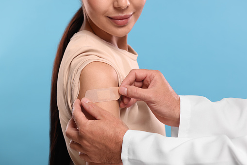 Doctor sticking plaster on woman's arm after vaccination against light blue background, closeup