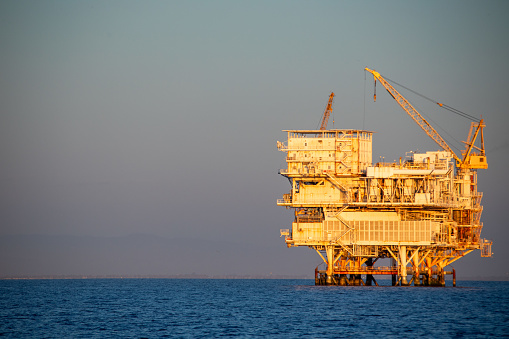 Oil Platform Gail during the golden hour in the pacific ocean off the coast of Ventura, Oxnard, Santa Barbara region between the coast and the Channel Islands National Park. Crane is loading supplies to an adjacent boat next to the rig / platform. 4HGX+2G