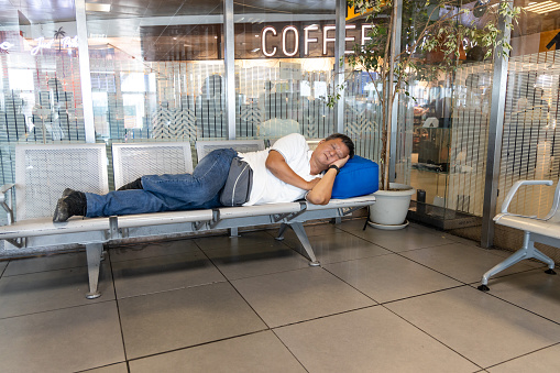 Tired Asian man sleeping flat on chairs at airport departure terminal waiting area while waiting for flight