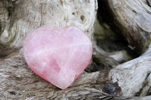A close up image of a beautifully shaped rose quartz crystal heart neatly resting a sun-bleached piece of driftwood.