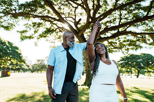 A vibrant and loving African American senior couple playfully dances in the shade of a tree while spending a relaxing afternoon together at the park during a vacation in Hawaii.
