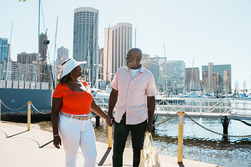 An active and loving African American senior couple affectionately hold hands and cheerfully talk while on a relaxing walk along a marina boardwalk with Honolulu, Hawaii skyline visible in the background.