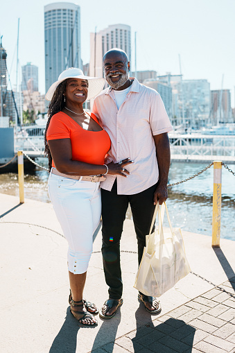 A vibrant African American senior couple affectionately embrace and smile directly at the camera while on a relaxing walk along a marina boardwalk with Honolulu, Hawaii skyline visible in the background.