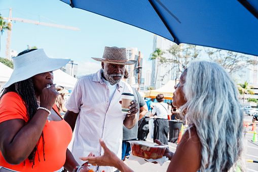 An active and adventurous African American senior couple try fresh tropical fruit and cheerfully chat with their local tour guide, a senior woman of Hawaiian decent, while at a farmer's market in Honolulu, Hawaii.