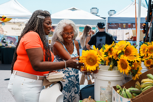 A vibrant and adventurous African American senior woman cheerfully talks with her local tour guide, a senior woman of Hawaiian and Chinese descent, while looking at fresh flowers at a farmer's market in Honolulu, Hawaii.