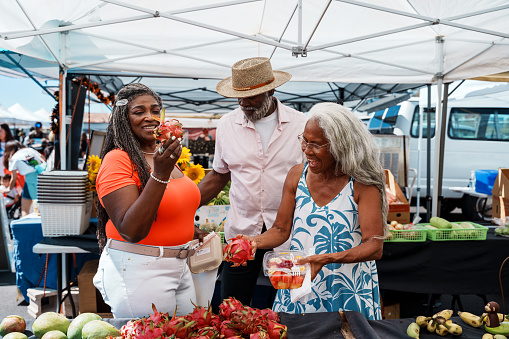 A vibrant and adventurous African American senior couple talk with their local tour guide, a senior woman of Hawaiian and Chinese descent, while looking at dragonfruit and other fresh tropical fruits for sale at a farmer's market during a tour of Honolulu, Hawaii.