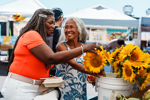 A vibrant and adventurous African American senior woman cheerfully talks with her local tour guide, a senior woman of Hawaiian and Chinese descent, while looking at fresh flowers at a farmer's market in Honolulu, Hawaii.