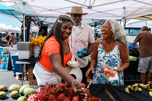 A vibrant and adventurous African American senior couple talk with their local tour guide, a senior woman of Hawaiian and Chinese descent, while looking at dragonfruit and other fresh tropical fruits for sale at a farmer's market during a tour of Honolulu, Hawaii.