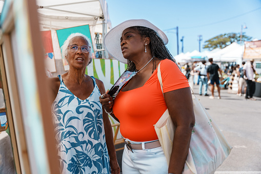 A senior woman of Hawaiian and Chinese descent, who is working as a local tour guide, talks with a senior woman of African American descent while at a farmer's market during a tour of Honolulu.