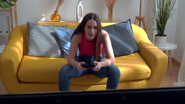 Young woman playing video games at home sitting on a yellow sofa