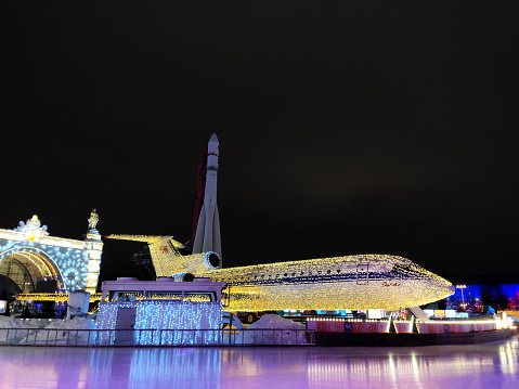 Moscow, Russia  December 23, 2023: Yak-42 aircraft in VDNKh park decorated with lights for Christmas and New Year celebration as part of the ice skating rink. Passenger plane monument with the space rocket Vostok in the background with illumination at night