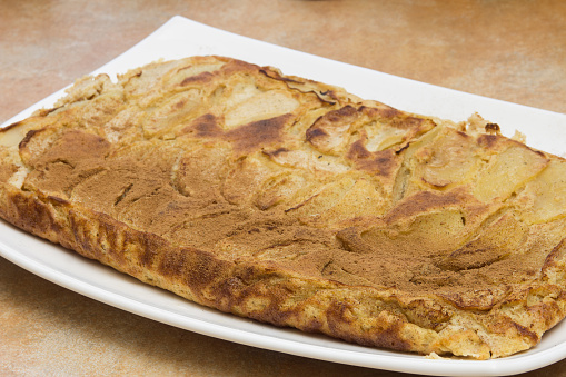 A close-up view of a golden brown and slightly crispy edge of a freshly baked apple pie with a light dusting of cinnamon on the overlapping layers of thinly sliced apples on a white rectangular plate.