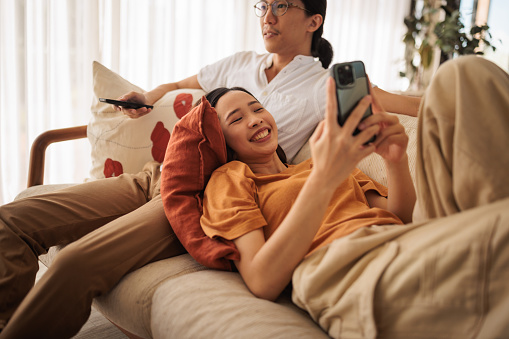 A smiling woman uses her phone while her husband watches TV in the living room.