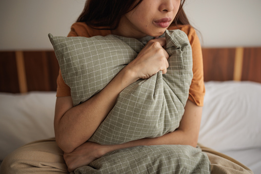 Young woman is sitting sadly on the bed, hugging a pillow.