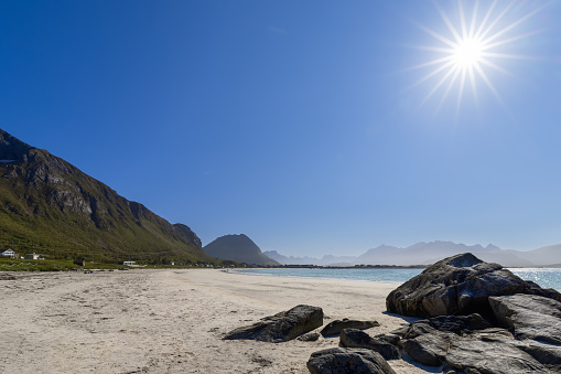 Sun rays piercing the blue sky at Jusnesvika Bay, foreground featuring dark, smooth rocks on Rambergstranda's white sand beach, with Flakstadoya's lush green slopes and hazy mountain silhouettes in the distance, in Lofoten, Norway