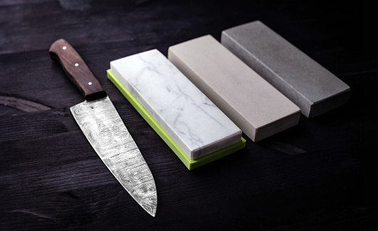 Big Kitchen Knife Rests On Kitchen Table, Accompanied By Set Of Sharpening Whetstones, All Against Black Background
